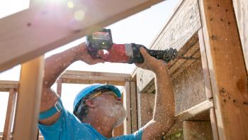 A volunteer wearing a blue Habitat shirt and hard hat holds a drill above his head while sawdust falls around him.