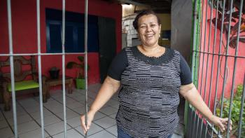 Brazilian woman smiling as she opens the gate to her pink house.