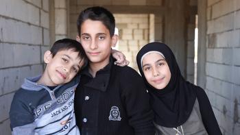 Two Syrian boys and a girl are showing in a concrete block hallway.