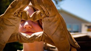 Hands in leather work gloves making a heart.  A woman's face can be seen through the heart.