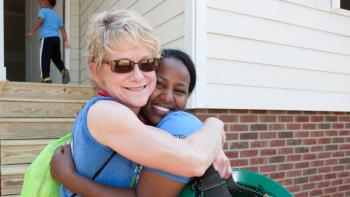 Two women hugging in front of a Habitat house with a little boy looking inside the front door.