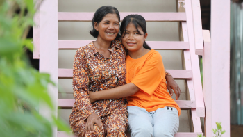 A Cambodian woman and her adoloescent daughter are smiling with their arms around one another sitting on the pink wooden stairs of their Habitat home.