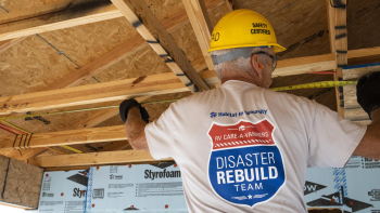 A man in a yellow hardhat that reads "safety certified" is shown from the back as he measures wood studs in a building under construction. The back of his shirt has a design saying "Habitat for Humanity RV Care-A-Vanners Disaster Rebuild Team"
