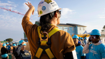 A woman wearing a hardhat and yellow safety gear has her back turned to the camera and her hand raised as she speaks to a group of people in blue volunteer shirts on a build site.