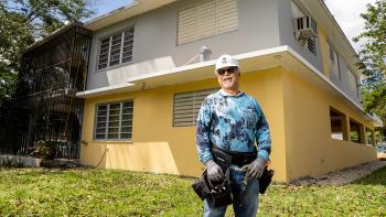 José with hard hat and tool belt in front of home repair site.