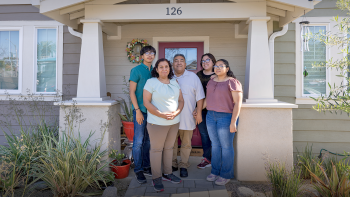 A mother and father pose standing together with their two young adult daughters and teenaged son on the fromt steps of their craftsman-style Habitat home.