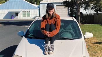 Girl in black ball cap, orange sweatshirt, leggings and tennis shoes sitting on the hood of a white car.