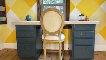 Blue desk with light wood top in front of a yellow wall with a chair in front. 