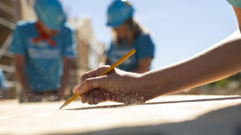 A close-up photo of a hand at a drawing board with two volunteers in blue shirts and hard hats blurred out in the background.