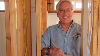 Cliff in a blue shirt, holding a pencil to paper as he stands in a partially constructed house.