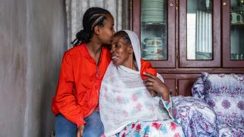 A mother sits in a white scarf and dress and her daughter in an orange shirt sits beside her and kisses her forehead.