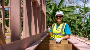 A woman in a bright blue T-shirt, hardhat and yellow vest gives a thumbs-up while standing between wooden slats of a Global Village house build. Palm trees are in the background.