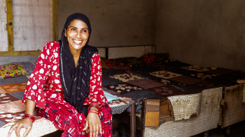 A woman sits in her home wearing a black head scarf and red shirt and pants, smiling from the camera.