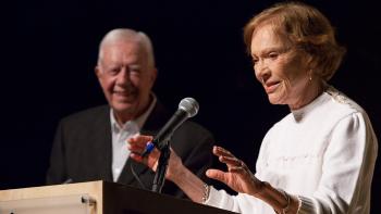 Mrs. Carter standing at a microphone on a podium with her hands raised as she talks to the audience. President Carter is standing to the side smiling at her.
