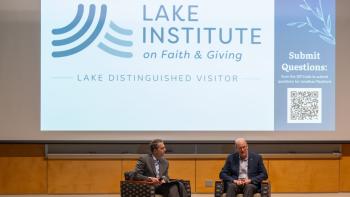 Jonathan Reckford and another man sitting onstage in chairs in front of an audience. In the background, a projector screen displays "Lake Institute on Faith & Giving" "Lake Distinguished Visitor" and a QR code. 