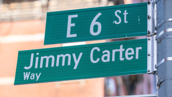 Close-up photo of street signs in the foreground and a brick building in the background. The street signs read "E 6 St" and "Jimmy Carter Way."
