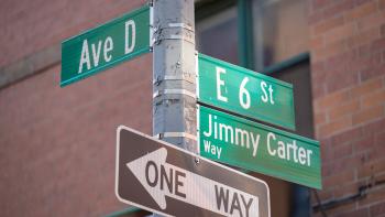 Street sign pole. Green street sign at top left reads "Ave D." On the right, green street signs read "E 6 St" and "Jimmy Carter Way." Below the other signs, a black and white arrow sign reads "ONE WAY."."