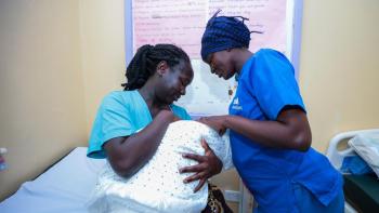 Kenyan nurse dressed in blue scrubs checking on a Kenyan mother and her new baby in a clinic.
