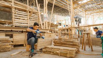 A man is sitting in warehouse surrounded by lumber and hammering a wooden pallet together.