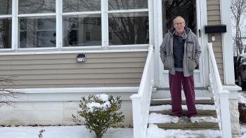 An older man in a jacket stands on the porch of his white house, surrounded by snow.