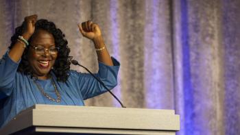 Black woman in a blue blouse celebrating with her hands in the air at a podium with HFHI's logo. 