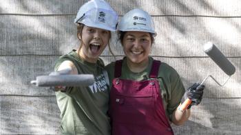 Two women with white hard hats and paint rollers against the side of a house.