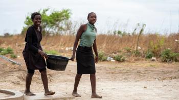 Two young ladies carry a bucket of water from a kiosk.