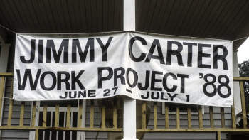 A large white banner hangs on the front porch of a home. The black text says "Jimmy Carter Work Project '88 June 27-July 1"