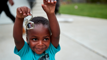 Toddler boy with arms up holding out a house-shaped keychain with key.