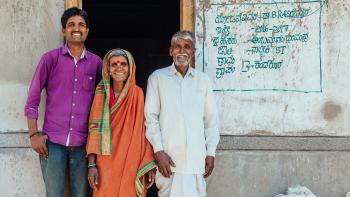 Indian family of three smile in front of their concrete home.