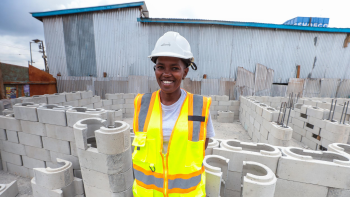 Woman in yellow safety vest and hard hat smiling and standing amongst concrete blocks at a construction site in Kenya