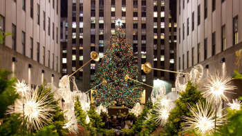 A very large decorated Christmas tree stands in Rockefeller Center plaza in NYC surrounded by decorative lights and angel figures holding golden trumpets.