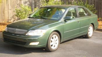 A dark green older model car in a parking space by a fence.