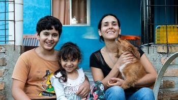 Patricia sits, smiling, with her two children and dog in front of a bright blue home.