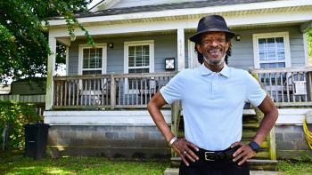 An older man in a fedora and polo shirt stands proudly in the front yard of his Habitat home in New Orleans.