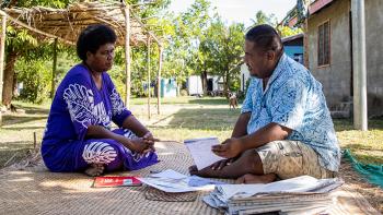 Two people in brightly colored clothing discussing paperwork while sitting on a straw mat outside with small concrete homes visible in background.