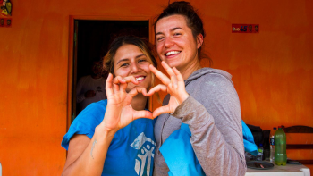 Two young women volunteers with Habitat shirts smile and pose with their hands making a heart shape.