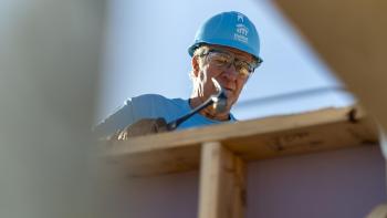 Volunteer in blue Habitat shirt and hard hat using a hammer on a rooftop.