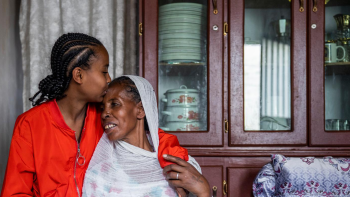 A mother in a white scarf and dress sits next to her daughter who is wearing an orange jacket. She kisses her mother's forehead.