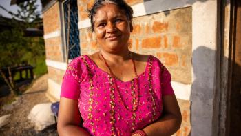 Niru stands outside her front door. She is wearing a pink top and the sun is behind her. She is smiling for the camera.