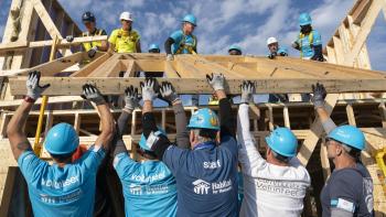 Habitat volunteers in blue shirts pulling and pushing up on lumber to assist in building a roof.
