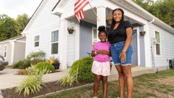 Mother and daughter smiling and standing in front of their newly built white house with an American flag hanging from it.