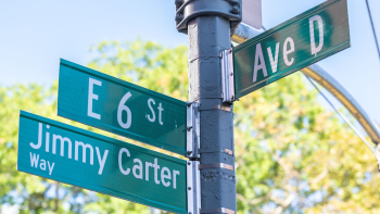 Low-angle photo of New York street signs at an intersection. The signs are green with white letters. On the left, the top sign reads "E 6 St" and the bottom sign reads "Jimmy Carter Way," while the sign on the right reads, "Ave D."