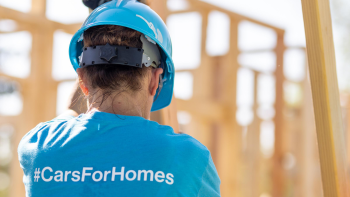 Habitat volunteer wearing blue hard hat and shirt that reads #CarsForHomes helps raise a wall on a build site.