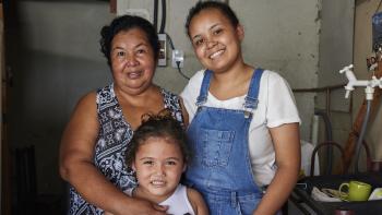 A mother embraces her two daughters in their home in an informal settlement, which has gray walls.