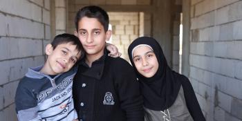 siblings from Syria living in Lebanon - two boys and a smiling girl wearing black scarf, new brick wall in the background