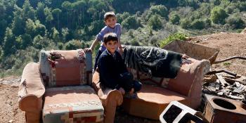Photo: two boys sitting on an old sofa outside. Mountains in the background.