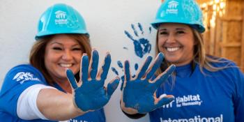 Two smiling women volunteers in hardhats hold up their hands which are painted blue.