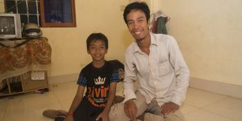 A man and child kneel on the floor of their home together, smiling.