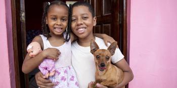 Young girl holding a doll with boy holding a dog in front of the front door to their home.